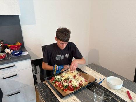 Eine Person sitzt an einem Tisch und bereitet eine Pizza auf einem Backblech zu. Eine Person sitzt an einem Tisch und bereitet eine Pizza auf einem Backblech zu.
