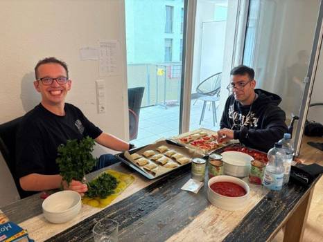 Zwei Personen sitzen an einem Tisch und pausieren bei der Zubereitung von Gerichten. Beide Personen lachen und schauen Richtung Kamera. Zwei Personen sitzen an einem Tisch und pausieren bei der Zubereitung von Gerichten. Beide Personen lachen und schauen Richtung Kamera.