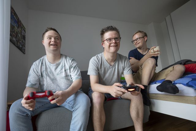 Drei Personen sitzen nebeneinander, zwei haben Handgeräte für eine Playstation in ihren Händen. Drei Personen sitzen nebeneinander, zwei haben Handgeräte für eine Playstation in ihren Händen.