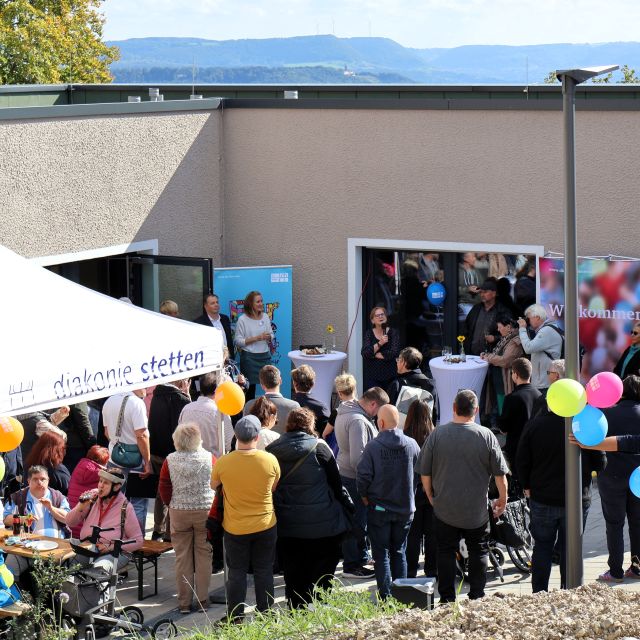 Bei der Einweihung des Wohnhauses in Göppingen-Hohenstaufen stehen und sitzen zahlreiche Personen vor dem Gebäude. Aufgebaut sind vor dem Gebäude Stehtische, ein Zelt und Banner der Diakonie Stetten. An einem Masten hängen drei bunte Luftballons. Bei der Einweihung des Wohnhauses in Göppingen-Hohenstaufen stehen und sitzen zahlreiche Personen vor dem Gebäude. Aufgebaut sind vor dem Gebäude Stehtische, ein Zelt und Banner der Diakonie Stetten. An einem Masten hängen drei bunte Luftballons.