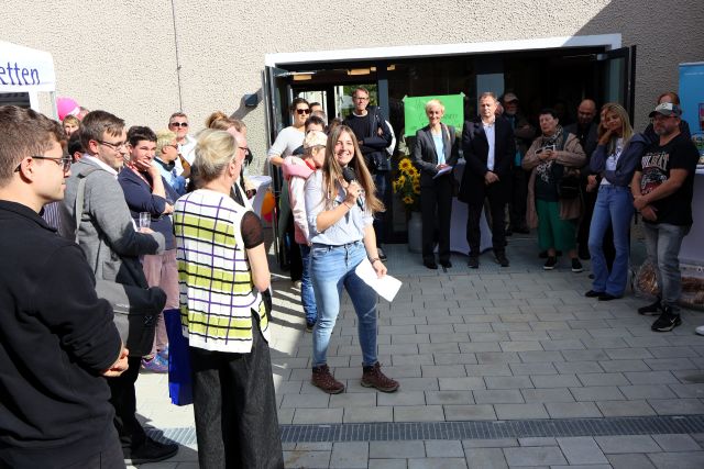 Eine Person spricht bei der Einweihung des Wohnhauses in ein Mikrofon, zahlreiche Personen hören zu. Eine Person spricht bei der Einweihung des Wohnhauses in ein Mikrofon, zahlreiche Personen hören zu.