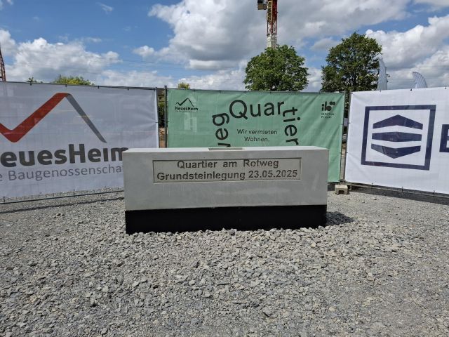 „Quartier am Rotweg – Grundsteinlegung am 23.05.2025“ ist auf einem Betonblock vor Bauzäunen mit Werbebannern zu lesen. „Quartier am Rotweg – Grundsteinlegung am 23.05.2025“ ist auf einem Betonblock vor Bauzäunen mit Werbebannern zu lesen.